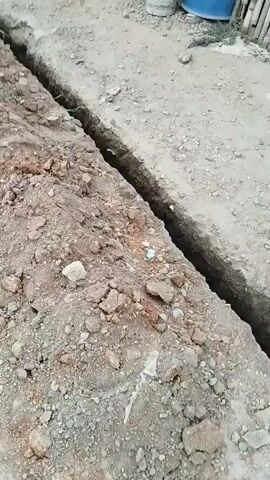 The video shows a long, narrow trench dug in the ground. The trench runs along the edge of a concrete or paved area. One side of the trench has a pile of excavated dirt and rocks. Blue plastic barrels are visible in the background. The camera moves slowly along the trench, providing a view of its length.
