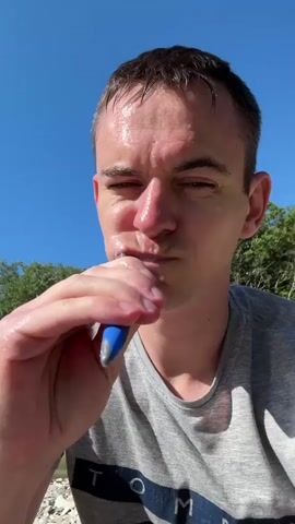 A man with wet, dark hair and a glistening face is seen in a close-up to mid-shot, brushing his teeth with a blue toothbrush. He is wearing a grey t-shirt with "TOMMY" visible on the front, likely a Tommy Hilfiger logo. He appears to be outdoors on a bright, sunny day, with a clear blue sky above. The man is squatting or sitting on a riverbank composed of small, light-colored pebbles and stones. Behind him, a shallow, clear river flows, and beyond that, green trees and hills or mountains are visible. He initially looks down, then up, and brings the toothbrush to his mouth, beginning to brush his teeth. Towards the end of the clip, he shows a slight smile with some toothpaste foam. The scene suggests an outdoor activity or camping trip in a natural, serene environment. The location is Shirokaya Schel, Russia.