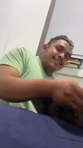 A man in a light green t-shirt and blue pants sits on what appears to be a bed. He is looking at the camera and talking, while holding a book with a blue and gold cover. Behind him, there is a white wall with a shelf that holds books and a water bottle. The man has a mustache and his hair is styled.