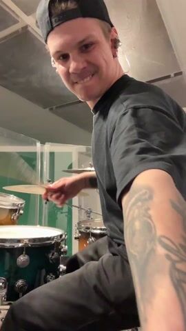 A young man wearing a black t-shirt and black baseball cap is playing a drum set in a recording studio. He is smiling at the camera as he plays. He has a tattoo on his left forearm. The drums are green and gold. The recording studio has acoustic panels on the walls and ceiling. There are keys hanging from his pants.