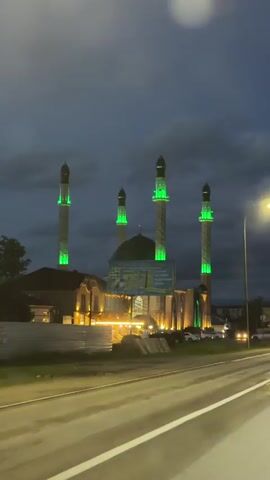 This video captures a nighttime view of a mosque with several tall minarets illuminated in bright green. The mosque features a large green dome and is surrounded by buildings with warm yellow lighting. The scene is viewed from a roadside, with a street and passing cars visible in the foreground. The sky is dark and cloudy, indicating it is nighttime. The overall impression is one of a well-lit and impressive religious structure against a dark, atmospheric backdrop.