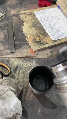 In a workshop setting, likely in Sokury, Russia, a person is pouring a dark liquid from a stainless steel thermos into a small black cup. The liquid, possibly coffee or tea, is cascading into the cup, creating a small splash. The thermos has a black cap that also serves as a drinking vessel. Nearby, a pair of orange-handled scissors and a crumpled piece of white cloth are visible on a textured workbench. In the background, a sheet of paper with handwritten text and a grid pattern rests on a rough piece of wood. A metal ruler is also present on the workbench. The overall scene suggests a moment of pause or preparation for work, with the focus on the act of pouring a hot beverage in a functional, potentially manual labor environment. The lighting appears to be indoor, with no indicators of time of day or weather.