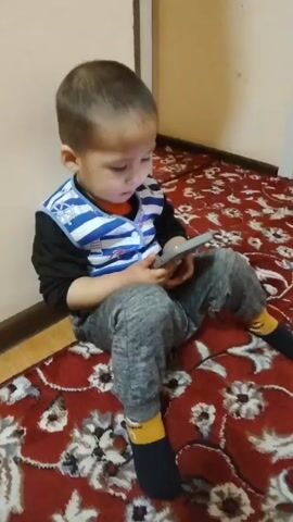 A young boy is sitting on a red and brown patterned rug, holding a cell phone in his hands and watching it intently. The boy is wearing a black long-sleeved shirt under a navy blue and white striped vest, gray sweatpants, and dark blue socks with yellow stripes at the ankles. He is sitting in a corner where a beige wall meets the floor. Some background music is playing.