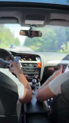 Two people sit in the front of a car, their arms leaning on the center console. Both individuals are wearing white shirts, and each is holding a smartphone. The person on the left is operating their phone, while the other person's phone is held stationary. The driver of the car is visible in the rearview mirror. The car is moving, as suggested by the passing trees visible through the windshield, and the sunroof is open.  The dashboard of the car, an Audi, is in view, showing the car's stereo, air vents, and gear shift. A pair of sunglasses sit atop the dashboard.