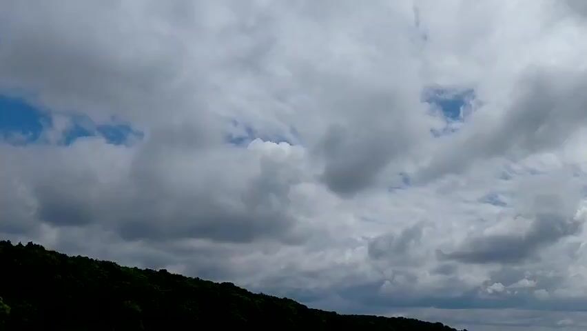 This video captures a panoramic view of a cloudy sky above a silhouetted forested hill. The clouds are dense and varied, with some areas of blue sky peeking through. The hill is covered in dark green foliage, and its outline slopes from left to right across the frame. The horizon line is visible in the distance, with hints of a flat landscape below the clouds. The lighting suggests an overcast day with diffused sunlight.