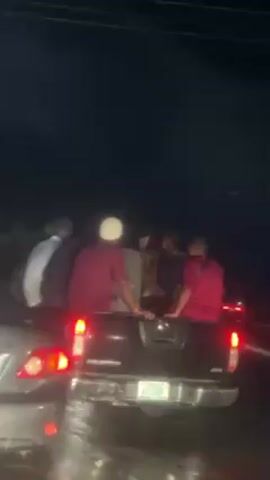 The video depicts a group of people riding in the back of a black pickup truck at night. The truck is on a wet road, likely after rain, as evidenced by the reflections and damp surface.  There are at least five people visible in the truck bed; they are wearing a mix of clothing including red shirts.  The license plate of the truck is partially visible. There is a car with illuminated tail lights in front of the truck, and a vehicle behind it. Overhead power lines are visible. The overall scene is dark due to the nighttime setting.