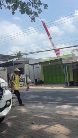 A man wearing a yellow shirt and black pants walks along a sidewalk next to a road. He is carrying a bucket in his right hand and has a backpack on his back. A red and white flag is attached to a pole and is positioned to his left, extending upwards into the sky. Several houses and buildings are visible behind him, as well as power lines. A white car is parked on the left side of the frame. The sky is blue with some clouds.