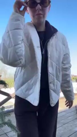 The video shows a woman standing on a wooden deck in what appears to be a backyard or outdoor area, likely in a suburban or rural setting. The weather seems sunny, with a clear blue sky overhead. The woman is wearing sunglasses, a light-colored quilted jacket, a dark-colored top with a collar, and dark pants. Her hair is styled in a bun. In the background, there's a wooden fence decorated with greenery. Beyond the fence, there is snow-covered ground, trees, and some houses in the distance. The woman adjusts her sunglasses, poses, turns to the side, and raises her arm with fingers in a "peace" sign. The camera angle is relatively low, shooting upwards toward the woman and the sky.