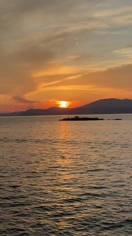 The video shows a scenic sunset over a body of water, likely an ocean or a large lake. The sun is positioned low on the horizon, with its light reflecting on the water's surface. The sky is filled with clouds, tinted in shades of orange, pink, and yellow due to the sunset. In the distance, there are silhouettes of mountains, adding depth to the landscape. A small, dark islet with a building on it lies between the viewer and the horizon, enhancing the visual interest of the scene. The water is calm with small ripples reflecting the light.
