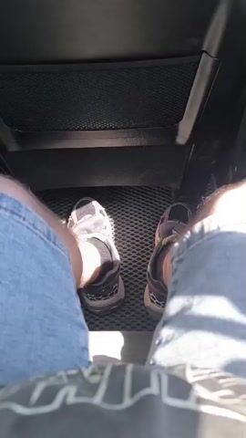 The video shows a first-person perspective of someone sitting in a car. The camera is pointed downwards at the person's feet, which are clad in sneakers and blue denim shorts. The feet are resting on the car's floor mat, which has a textured pattern. Sunlight is visible, casting shadows on the floor. The background shows the back of a car seat with a mesh pocket. The person's legs are positioned slightly apart, and their knees are bent. The view of the feet and legs shifts slightly as the camera or the person moves.