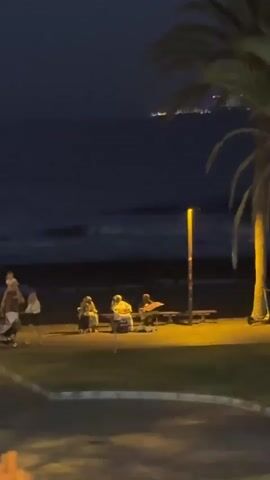 The video shows a nighttime scene at a beach. There is a dark ocean with waves in the background. On the shore, there is a sandy area with some people sitting at a picnic table under a lamppost. There are also some palm trees and grass in the foreground. In the distance, there are lights from a city. The video appears to be taken from a slightly elevated position across a road or walkway. The lighting suggests it is dusk or night.