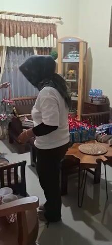 The video shows two people in a room. One person, wearing a white t-shirt with a floral design and a black headscarf, is standing and facing away from the camera, looking downwards. They appear to be laughing or smiling. The other person, wearing a grey dress and a black headscarf, is standing to the left and facing away from the camera, towards a shelf filled with items. In the foreground, there are chairs and a coffee table. The room appears to be a living room, with curtains and furniture visible. There are several gift bags arranged on a coffee table in the middle of the room.