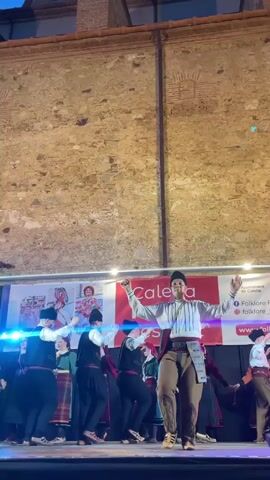 The media file shows a group of people performing a traditional dance on a stage. The performers are dressed in colorful, traditional folk costumes. The person in the foreground, a young male, is the most prominent. He is wearing a white shirt with intricate embroidery, a sash, brown pants with decorative stitching, and a black cap. His arms are raised and outstretched, and he appears to be in the middle of a dynamic dance move, with one leg bent and the other extended.

Behind him, several other dancers are visible. The women are wearing long, patterned skirts and blouses, while the men are dressed in vests, shirts, and dark pants, also adorned with sashes and belts. Some of the dancers are holding instruments or props, though they are not clearly discernible. The backdrop features a banner with images of people in traditional attire and text.

The scene is set outdoors at what appears to be an evening event, indicated by the stage lighting. The performance is taking place in front of a textured, rustic wall, likely made of brick or stone. There are banners with visible text, including "Ajuntament de Calella" (Calella City Council) and "folklore" along with social media icons and website URLs. The overall atmosphere seems festive and engaging, with the dancers displaying energy and precision in their movements. The lighting suggests it is dusk or early evening.