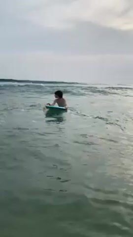 A young boy is seen in waist-deep ocean water, bodyboarding. He is light-skinned with dark hair and wearing a dark wristband on his left wrist. He paddles, catches a small wave, and raises his left hand in a wave, before continuing to lie on the board. The board is light blue and green. The ocean is green with visible waves in the distance under an overcast sky. Other people are vaguely visible in the distance.