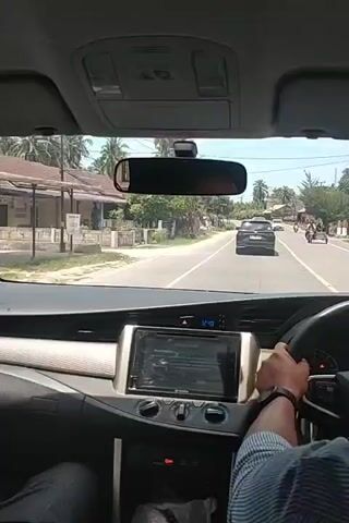 From the perspective of a driver inside a car, the vehicle is driving down a paved road in a tropical environment. Houses and lush foliage line the sides of the street, with tall palm trees swaying in the breeze.  Ahead, two other cars and a motorcycle can be seen on the road. The interior of the car is visible: a dashboard with a display screen showing 12:42, a rear-view mirror, and a person’s arm holding the steering wheel. The person is wearing a blue shirt. The scene is captured on a bright, sunny day.
