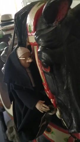 A close-up shot shows a small, elderly doll with pale skin, glasses, and a prominent nose, dressed in a black cloak and a white cap, sitting on a miniature black carousel horse with red and white accents. The doll's hands are placed on the horse's neck. In the background, out of focus, a room with various objects like hats and papers is visible. The camera slowly zooms out, revealing more of the horse and the cluttered background.