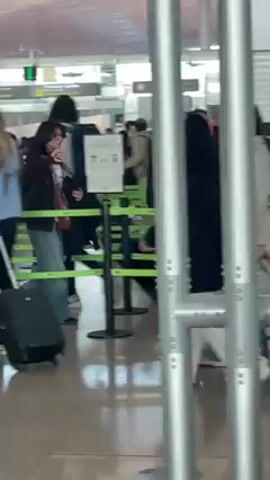 The video shows an indoor scene, presumably an airport security line or checkpoint. Several people are walking in the same direction behind green and white barriers. Some have backpacks and rolling luggage. The camera is positioned behind a metal railing, creating a vertical bar effect in the foreground. Signage in Spanish is visible in the background, including the phrase "Control de seguridad." The overall lighting appears bright. The video is a moving perspective as the camera holder follows people walking in the same direction.