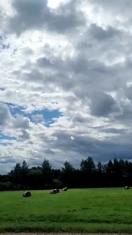 The video shows a view from a vehicle moving past a field with several hay bales. The sky is filled with dramatic, grey and white clouds, with patches of blue visible. A dark line of trees runs along the horizon behind the field. The field itself is bright green, and the hay bales are round and dark brown. The vehicle appears to be on a paved road, and there is a railing visible in the foreground.