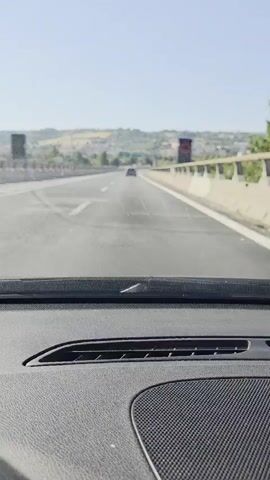 The video shows a front-facing view from inside a car driving on a multi-lane highway on a sunny day. The road is clear with white lane markings. In the distance, other cars are visible. The highway is flanked by concrete barriers and a sloped, grassy terrain with some buildings and trees. The sky is clear and blue. The car's dashboard is visible at the bottom of the frame. The video captures the movement of the car as it travels forward, with the landscape slowly changing in the background.