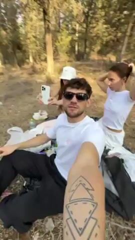 The video begins with a selfie-style shot of a young man wearing sunglasses and a white t-shirt. He is sitting on the ground with two young women. One woman is wearing a white bucket hat and light pink clothing, holding a phone. The other woman is wearing a white top and pulling her hair up. They are sitting on a blanket surrounded by trees. The camera then pans around to show the forest setting with tall pine trees and a dirt ground covered in pine needles.