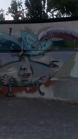 The media shows a section of a colorful mural painted on a concrete wall. The mural depicts a surreal and abstract composition with elements that resemble distorted faces, fruits, and fantastical creatures. In the foreground, a small, white mouse with pink ears is painted, looking directly at the viewer. Behind the mouse, a large, distorted human face is integrated into the artwork, with wide-open eyes and a mouth that appears to be mid-expression. Above this face, there are stylized representations of a pear and an apple, with the apple featuring a ladybug on its surface. To the right of the apple, a slice of watermelon is visible. Further to the left, sketches of what appear to be a robot and an alien-like figure are drawn on the wall, almost as preliminary studies for the main mural. The overall style is a blend of realism and cartoonish elements, creating a dreamlike or unsettling atmosphere.

The setting appears to be outdoors, indicated by the presence of trees and the natural lighting, suggesting it might be daytime. The ground in front of the mural is paved with asphalt, and there are some small plants growing near the base of the wall. The concrete wall itself has areas where the paint is chipped or worn, revealing the underlying material. The sky is visible at the top of the frame, and the light suggests it could be late afternoon or early evening. There are no visible signs of text within the mural itself. The artwork appears to be located in an urban environment, possibly a public space or an alleyway, given the graffiti style and the presence of the wall. The combination of fantastical imagery and the raw urban setting creates a striking visual contrast.