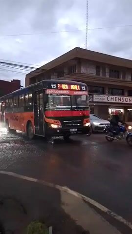 The camera follows an orange and black city bus labeled “MAGNO” as it turns right onto a rain-slicked street. The bus has the number 145 on its front and sides and lists San Lorenzo and Asuncion as destinations. A silver car is visible in the background, and a person on a motorcycle passes on the right. Buildings are seen in the background, one labeled “CHENSON.” The sky is overcast.