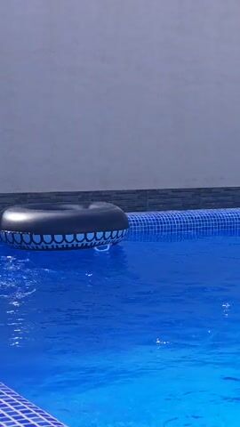 A black inflatable pool float with a blue and white tooth-like pattern is floating in a bright blue swimming pool. The pool is partially visible, with a tiled edge in the foreground. The water is clear and appears still, with some ripples on the surface. The background is a neutral gray wall. The lighting suggests it is a sunny day.