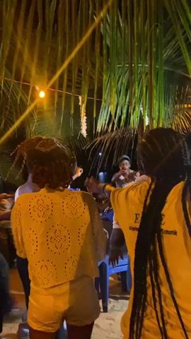 A group of people are gathered under a palm tree at night. The people are of African descent. Many are dancing around a person seated on a blue chair, seemingly celebrating them. One person in the crowd is wearing a bright yellow shirt with the inscription "ESAE". A string of white decorations hangs from the palm tree above the group. A warm light source illuminates the scene from the left.