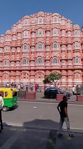 The video shows a daytime view of the Hawa Mahal in Jaipur, India. The building is a five-story structure made of red and pink sandstone, with a multitude of small windows and balconies decorated with intricate carvings. People are walking along the street in front of the building, and there is a small tree in the foreground. A man is walking across the foreground.  The sky is clear and blue.