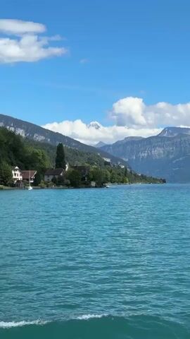 The video begins with a view of a vibrant blue lake with rolling waves. In the distance, a lush green coastline is visible, dotted with houses and trees. Towering mountains, some with snow-capped peaks, form the backdrop. A sailboat can be seen in the middle ground on the lake. The camera pans slightly to the right, revealing the side of a boat, including a railing. The sky is a bright blue with scattered white clouds.