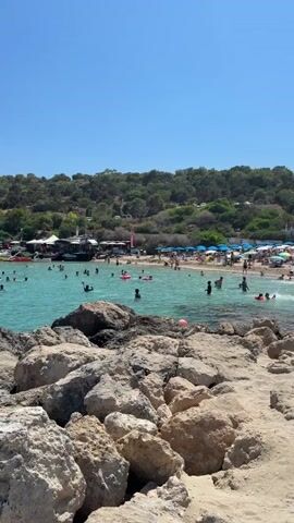 The video shows a crowded beach on a sunny day. The water is a clear turquoise color, and many people are swimming and playing in it. There are also some boats and inflatable toys in the water. The beach is lined with rocky outcrops in the foreground, and a green, tree-covered hillside in the background. There are several buildings and umbrellas visible on the beach, suggesting a resort or tourist area. The sky is clear and blue.