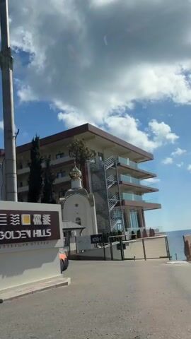 The video appears to be shot from inside a vehicle, looking out at a modern hotel or resort building. The building is multi-story with a light-colored facade and brown accents. There are several cars parked in front of the building, including a white van and two sedans, one of which is silver. Trees and dense green foliage partially obscure the view of the building and the cars. In the foreground, parts of the vehicle from which the video is being recorded are visible, including a side mirror and what looks like a hand resting on the car door. The sky is blue with scattered white clouds. The overall impression is a sunny day at a lodging establishment.