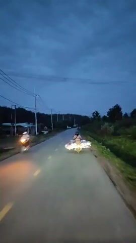 A motorcyclist rides down a road at dusk. The motorcyclist is carrying a large, colorful load of goods strapped to the back of the motorcycle. The road is paved and has yellow lane markings. There are power lines and trees along the side of the road. The sky is dark blue and cloudy. The camera is mounted on another vehicle, also moving on the road.