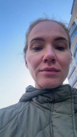 A fair-skinned woman with blue eyes and light blonde hair stands outside, wearing a green hooded jacket. The background shows a street with parked cars and buildings under a clear sky. The camera is positioned closely, focusing on her face.  The woman is looking directly at the camera with a neutral expression.