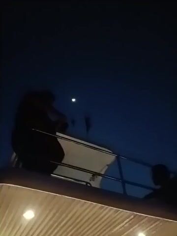 The video shows a dark scene at night. Two individuals are seen, primarily in silhouette, standing on what appears to be a balcony or elevated structure with railings. The sky above is a deep blue, and a crescent moon is visible. The camera pans slightly, revealing one of the individuals up close. They have glasses. The sound of wind is prominent throughout the video.
