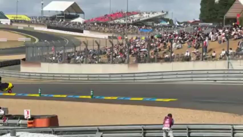 Here's a forensic description of the video:

**Scene:** The video captures a segment of a race track, likely a professional motorsports event.

**Environment:** The weather appears to be clear and sunny. There are large crowds of spectators lining the track, visible in stands and on grassy embankments. In the background, there are large temporary structures such as tents or canopies.

**Objects:**
*   A race car speeds past the camera. The car is primarily yellow and blue with a complex, abstract pattern. The car number is visible, reading "311".
*   The race track itself features multiple lanes, with painted track boundaries. There is a retaining wall alongside the track, with sponsor banners advertising Cadillac. A chain-link fence separates the track from the spectators.
*   There's a sign with the number "1" visible in the foreground.
*   Various pieces of trackside infrastructure are visible, including posts and small structures.

**People:**
*   Numerous spectators are present in the grandstands and along the side of the track.
*   One person is seen holding a camera, presumably photographing or filming the race.

**Actions:**
*   The race car is speeding along the track.
*   Spectators are watching the race.
*   A photographer is actively taking pictures.

**Interactions:**
*   The race car interacts with the track.
*   Spectators are interacting with the event by watching.
*   The photographer is documenting the event.