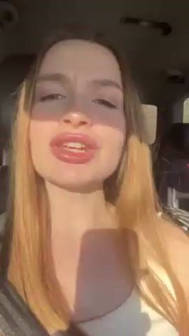 A young woman with long, light brown hair and bright red nails is seated in a car. She is looking directly at the camera and talking, gesturing with her hands. Sunlight illuminates her face from one side. The interior of the car is visible in the background, including the headrest of a car seat.