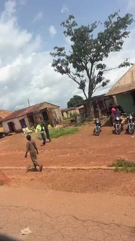 The video shows a street scene in what appears to be a rural or developing area. There are several buildings with corrugated metal roofs, some appearing to be shops or residences. The ground is a reddish dirt. In the foreground, a paved road is visible. There are a few vehicles, including a car and a motorcycle. Several people are present, some walking and some standing near the buildings. There are also some piles of goods or materials outside the shops. The sky is cloudy. The camera appears to be mounted on a moving vehicle, as the scene is slightly shaky and pans across the environment.