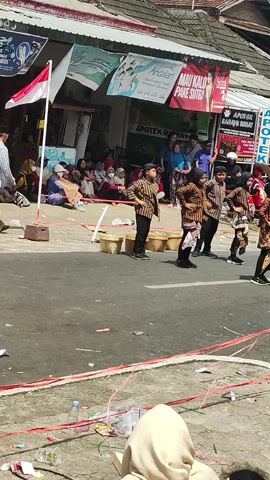 A group of children in traditional clothing are performing a dance on a paved road. They are wearing matching patterned shirts and sarongs. There are several wicker baskets placed on the ground in front of them. In the background, there are buildings with signs that appear to be shops, including one that says "APOTEK" which means pharmacy. A crowd of people are watching the performance from the sidewalk and behind a rope barrier. A flag, possibly the Indonesian flag given the red and white colors, is visible on a pole. The scene appears to be an outdoor event or festival.