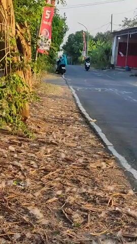 Two motorcyclists are riding on a paved road. The road is bordered by dry vegetation on the left and buildings on the right. The sky is clear and the sun is shining. The first motorcyclist is wearing a blue hijab and is stopped on the side of the road. The second motorcyclist is wearing a black jacket and a helmet and is riding a white scooter. There is a red and white flag on a pole on the left side of the road. There are drawings on the road in white paint.