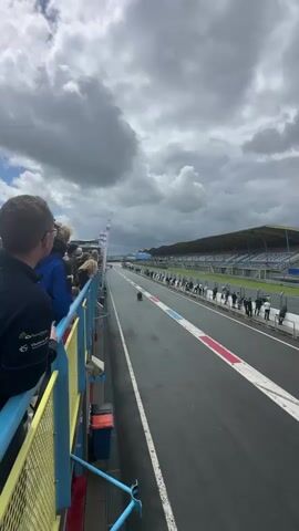 The video shows a race car driving on a race track. There are spectators on the left side of the track, looking towards the race. The track is wet, and the sky is cloudy. The race track has blue and red markings.