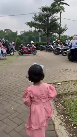 The video shows a partially cloudy day at an outdoor venue. The first shot focuses on a young girl in a pink dress and headband walking towards a motorcycle parking area. People mill around the parking area and motorcycles are parked closely. The video then shifts to show a wooden seating structure resembling a sliced barrel. Three people are seated inside at a table. The video pulls back to reveal another similar seating structure. Palm trees are present and the landscape includes trees and mountains in the background. The sky is overcast.