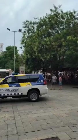 A police officer stands near a white and blue patrol vehicle with lights flashing. Across a paved plaza, two individuals in red and orange uniforms with red crosses on their vests stand observing. A building in the background is obscured by thick white smoke. Several trees are visible in the middle ground. A yellow vehicle and what appears to be a stage setup are also present in the background.