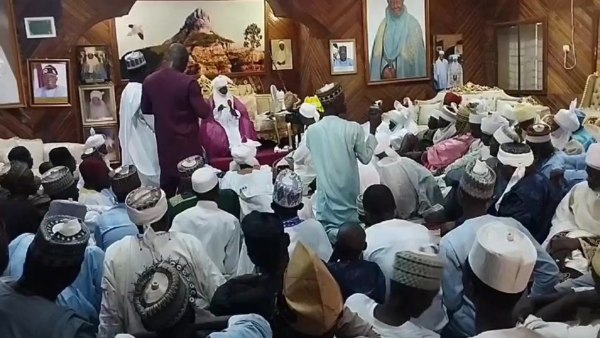 The video shows a large room, likely a meeting or audience hall, with wood-paneled walls adorned with framed portraits. A crowd of men, mostly wearing traditional Islamic attire, including robes and caps, are seated on the floor. At the front of the room, a man wearing a bright pink robe and a white cap sits on a raised platform or chair, suggesting a position of authority or importance. Several individuals stand around him. The atmosphere appears formal, possibly a gathering for an address or ceremony.