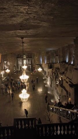 This image depicts a vast, dimly lit underground chamber, characteristic of the Wieliczka Salt Mine in Poland. The scene is dominated by several large, ornate chandeliers, which are the primary light source, casting a warm glow across the cavernous space.

Numerous people are visible on the chamber floor, clustered in groups, appearing to be tourists or visitors exploring the site. Their reflections can be faintly seen on the polished or wet surface of the floor. Some individuals are also positioned on a raised balcony or platform along the right side, looking down into the main area.

The walls of the chamber are rough and textured, likely carved from salt, with various architectural details and reliefs visible under spotlighting. On the left side of the frame, a long, sloped wooden ramp or staircase extends downwards, adding to the sense of depth. A sturdy wooden support structure or barrier is also visible on the right. The overall atmosphere is one of grandeur and historical significance, with the bright chandeliers providing a striking contrast to the dark, cavernous surroundings. No discernible text is visible in the image.