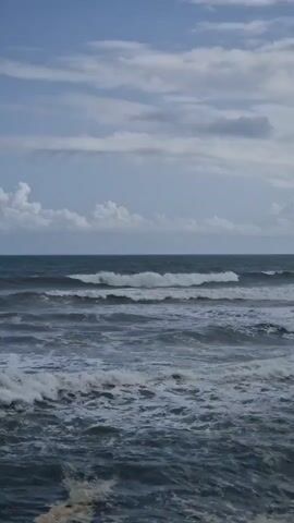 The video shows a wide shot of an open ocean under a partly cloudy sky. Gentle waves roll towards the shore, breaking into white foam. The horizon line is distinct, separating the sea from the sky. The cloud cover is scattered with patches of blue sky visible. The sea appears choppy with varying shades of blue.
