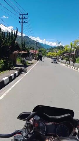 The video begins with a first-person view from a motorcycle. The rider is on a paved road in a town or village. To the left, there are houses and vegetation, with a steep, mountainous backdrop. Utility poles and wires run along the road. On the right side of the road, there are palm trees, streetlights, and businesses. Several vehicles are visible on the road, including cars and a truck. The sky is clear and blue. The camera pans slightly to the right, showing more of the streetscape and the surrounding mountains. In the foreground, the motorcycle's handlebars and dashboard are visible. The video is a short clip, likely captured while riding the motorcycle.