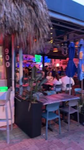 The video pans across an outdoor dining area of a restaurant situated along a paved walkway. Several tables with chairs are visible, some occupied by diners. The restaurant's exterior features a covered area with awnings, string lights, and an American flag flying from a pole. Menus are displayed on stands outside the restaurant. The walkway is lined with palm trees and other vegetation. People are walking along the walkway, suggesting it's a public thoroughfare. The overall atmosphere appears to be that of a lively evening scene at a beachside or boardwalk establishment.