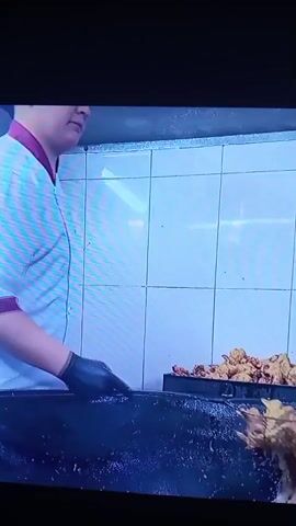 A person wearing a white chef's coat and black gloves is cooking and serving food. In the first few seconds, the person is using tongs to flip some fried food in a pan. Then, the person is seen serving a dish with what appears to be shredded vegetables and meat into a paper boat-shaped container, using their gloved hands to mix the food. The scene is set in a commercial kitchen with tiled walls and stainless steel surfaces. There are other containers of food visible in the background.