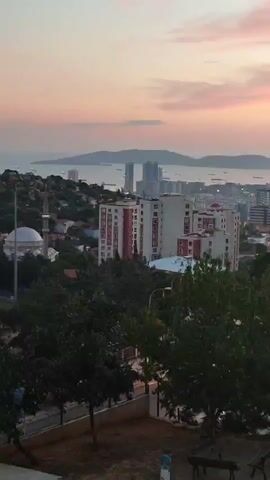 A high-angle, wide shot captures a city skyline at dusk. In the foreground, trees with dark green leaves frame the scene. A pathway with a fence runs along the bottom, leading to a park area with two benches, one of which is occupied by a person. In the midground, several multi-story apartment buildings dominate the view, with one prominent building featuring a red and white striped facade. To the left, a white domed building with a minaret, likely a mosque, is visible amongst smaller structures and trees. The background reveals a vast body of water, possibly a bay or sea, dotted with ships. Across the water, a distant city skyline rises, characterized by modern high-rise buildings and an island landscape. The sky above is painted with soft pink and orange hues, indicative of sunset, with wispy clouds scattered across it. The overall atmosphere is tranquil and serene.